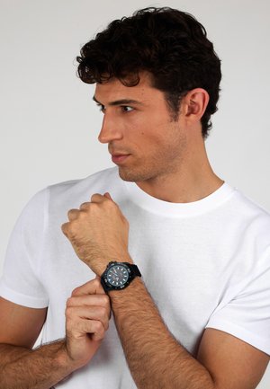Orologio analogico nero con cinturino in gomma texturizzata, cassa rotonda, quadrante ampio e indicatori bianchi a contrasto. Indossato su un polso, mostra le mani che lo regolano.