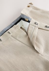 Pantalon beige avec ceinture partiellement ouverte montrant le bouton intérieur, les agrafes, la fermeture éclair et les détails de la doublure en tissu.