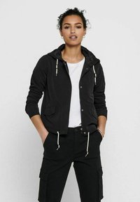 Giacca leggera nera con cappuccio, chiusura con zip, due tasche frontali e dettagli a cordino. Abbinata a una camicia bianca e pantaloni neri.