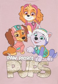 Koszulka Paw Patrol z trzema kreskówkowymi psami na jasnoróżowym tle. Psy noszą kolorowe stroje z wyróżniającymi się rysami twarzy. Napis „PUPS” w brokacie.