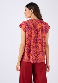Blusa floreale arancione con motivi a foglia bordeaux, maniche corte e vestibilità rilassata. Tessuto morbido con una finitura leggermente strutturata.