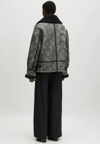 Graue Lammfelljacke mit schwarzem Kragen und Akzenten, die eine kastige Passform, eine weiche Textur und sichtbare Nahtdetails aufweist. Kombiniert mit lockeren schwarzen Hosen.