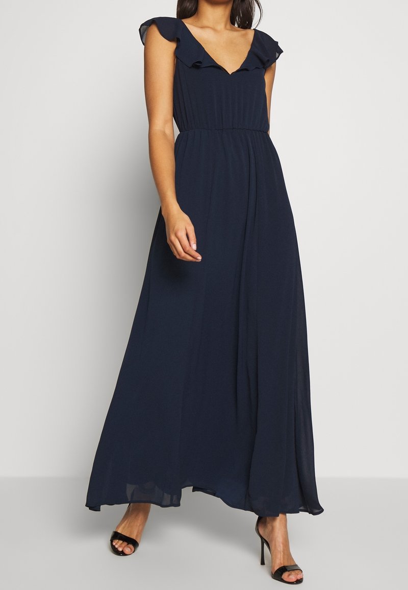 Femme portant une robe longue sans manches bleu marine avec un col à volants et des sandales à talons noires, debout devant un fond uni.