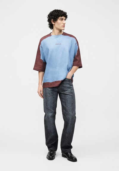 Νεαρός άνδρας με σγουρά μαλλιά, φορώντας φαρδιά πουκάμισα σε μπλε και καφέ χρώματα, παντελόνι με λεοπάρ σχέδιο και μαύρα επίσημα παπούτσια, όρθιος.