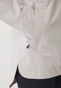 Helle graue wasserdichte Jacke mit einer Reißverschlusstasche, Seitennähten und dezentem Stickerei nahe dem Bündchen. Verfügt über einen leicht geschwungenen Saum.