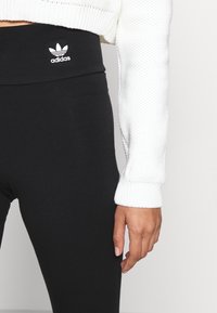 Svarta leggings i stretchmaterial med hög midja; har en vit Adidas-logotyp. Åtföljs av en vit strukturerad tröja.