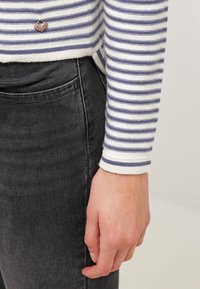 Mano che riposa accanto a jeans in denim neri, indossando un top a maniche lunghe bianco e blu a righe con un piccolo ciondolo a forma di cuore vicino all'orlo.