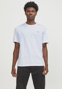 White cotton t-shirt with short sleeves, crew neck, and a small black logo on the left chest. Paired with dark jeans.