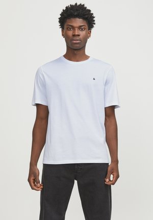 White cotton t-shirt with short sleeves, crew neck, and a small black logo on the left chest. Paired with dark jeans.