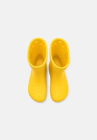 Une paire de bottes de pluie en caoutchouc jaunes vues de dessus sur un fond blanc, avec des trous de ventilation près du haut.