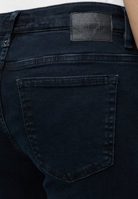 Gros plan sur la poche arrière d’un jean en denim bleu foncé avec des bords surpiqués et un écusson de marque en cuir noir portant la mention « DRYKORN » sur la ceinture.