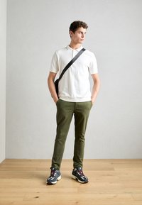 Hvid polo shirt, olivengrønne bukser, multikolorede sneakers. Modellen poserer med en sort crossbody taske foran en neutral baggrund.