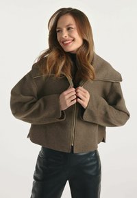 Brązowa krótka kurtka z szerokim kołnierzem i zapięciem na zamek; wykonana z miękkiego materiału; model ma na sobie dopasowane czarne spodnie; gładka tekstura i prosty design.