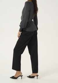 Graue Bluse mit langen Ärmeln und Taillengürtel, kombiniert mit schwarzen, weit geschnittenen Hosen. Schwarze, verzierte Mules mit Absatz vervollständigen den Look.