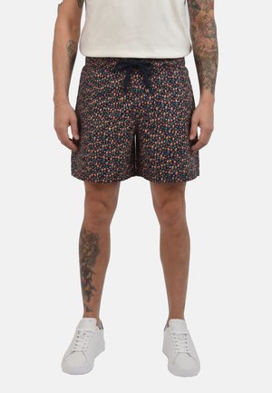 Uomo che indossa pantaloncini blu navy con piccolo motivo floreale multicolore, scarpe da ginnastica bianche e t-shirt bianca, in piedi con le mani rilassate.