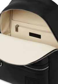 Sac à dos noir avec un extérieur lisse, un intérieur beige clair et une poche zippée. Dispose d'une fermeture éclair argentée et d'un détail de logo texturé.