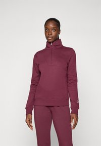 Bordeauxfarbener Pullover mit hohem Kragen und halbem Reißverschluss, gefertigt aus einem weichen Stoff. Besitzt eine glatte Textur und eine taillierte Passform, mit einem Logo auf dem Ärmel.