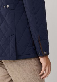 Marineblau gesteppte Jacke mit diagonaler Naht, braunen Akzenten an den Bündchen und Taschen, silbernen Druckknopfdetails und einer glatten Textur.