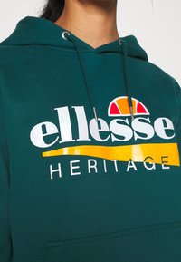 Personne portant un sweat à capuche vert avec le logo blanc « ellesse HERITAGE » et un motif semi-circulaire orange et rouge sur la poitrine.