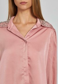 Rosa Satinbluse mit Knopfverschluss und verziertem Kragen. Verfügt über glänzendeDetails an den Schultern, eine glatte Textur und einen entspannten Schnitt.