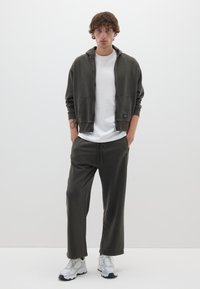 Dunkelgrüner Reißverschluss-Hoodie mit Fronttaschen, kombiniert mit einem hellgrauen T-Shirt und lässigen, dunkelgrauen Hosen. Weiße Sneakers mit grauen Akzenten.