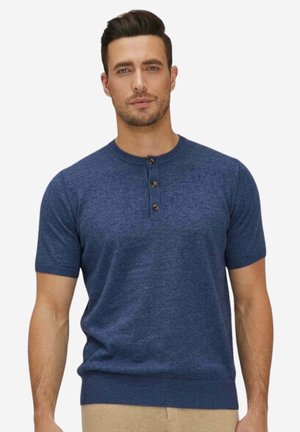 Mann trägt ein blaues Henley-Shirt mit kurzen Ärmeln und drei Knöpfen sowie beige Hosen, schaut nach vorne vor einfarbigem Hintergrund.