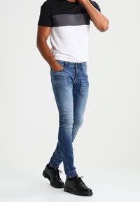 Camiseta a rayas en blanco y negro, jeans ajustados azules y zapatos negros con cordones. La camiseta tiene un cuello redondo y mangas cortas. Los jeans tienen una textura deslavada.