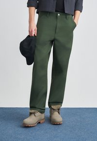 Πράσινο βαμβακερό παντελόνι με ίσια γραμμή, με τσέπες, κλείσιμο με κουμπιά και φορεμένο με μπεζ μπότες και μαύρο καπέλο.