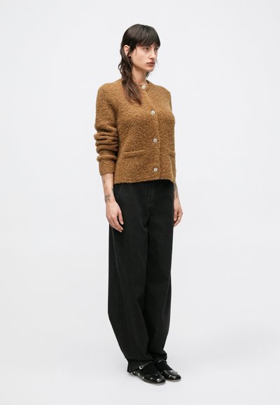Brązowy, miękki sweter na guziki z okrągłym dekoltem, pięcioma guzikami i dwoma przednimi kieszeniami, zestawiony z luźnymi czarnymi spodniami i czarnymi butami wsuwanymi.