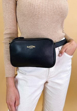 Black leather fanny pack with a textured finish, gold logo, rectangular shape, zip closure, worn at the waist over white pants.
