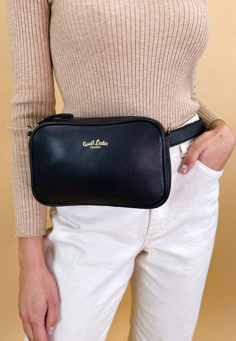 Marsupio in pelle nera con finitura testurizzata, logo dorato, forma rettangolare, chiusura con zip, indossato in vita sopra pantaloni bianchi.