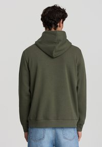 Uomo con i capelli castani ricci e corti, che indossa una felpa verde oliva e jeans azzurri chiari, rivolto di spalle su uno sfondo chiaro e uniforme.