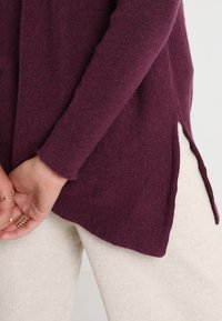 Pull à manches longues, d'un profond bordeaux avec une finition texturée et de hautes fentes latérales, associé à un pantalon texturé de couleur crème.