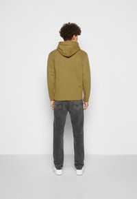Felpa con cappuccio verde oliva dal taglio rilassato, abbinata a jeans grigi scuri e sneaker bianche, vista da dietro su uno sfondo neutro.