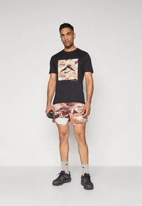 Mustast t-särgist, millel on mägipilt, maatoonides mustrilised lühikesed püksid, hallid sokid ja mustad spordijalatsid. Hoides musta veepudelit.