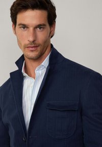 Blazer navy con un acabado texturizado, que presenta un cuello con muesca, cierre de un botón y un bolsillo en el pecho. Lleva puesto sobre una camisa de rayas clara.