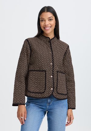 Femme souriante portant une veste marron à motifs boutonnée avec de grandes poches avant et un jean bleu sur un fond clair uni.