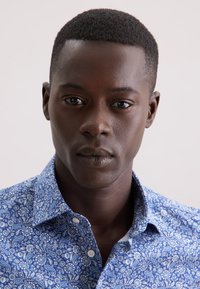 Giovane uomo di colore con capelli ricci corti, che indossa una camicia a maniche lunghe con motivo paisley blu, che guarda direttamente verso la macchina fotografica.