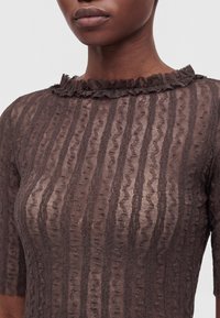 Gros plan d'un top en dentelle sombre et transparente avec des rayures verticales texturées et un col à volants porté par une personne à la peau de ton moyen-foncé.