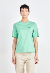 Camiseta verde menta de algodón, con cuello redondo, mangas cortas y un pequeño logo en el pecho. Combinada con pantalones de color claro.
