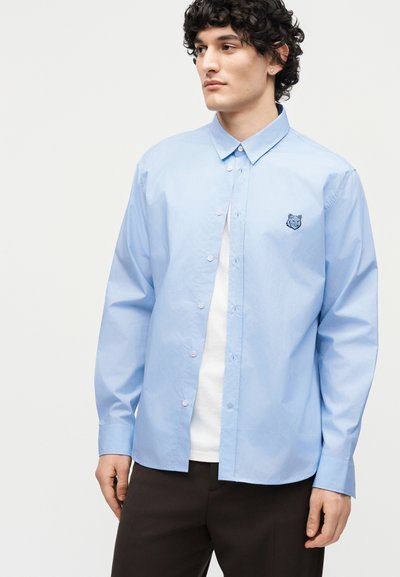 Άνδρας με κατσαρά μαλλιά που φοράει γαλάζιο πουκάμισο με κουμπιά και λογότυπο κεφαλής λύκου στο στήθος, λευκό μπλουζάκι και σκούρο παντελόνι.