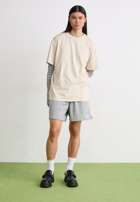 Tricou oversized bej stratificat peste un hanorac raiat cu mânecă lungă, pantaloni scurți gri, clogs negre și șosete albe stând pe un covor verde.