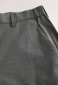 Gros plan sur un pantalon en tissu gris foncé montrant la boucle de ceinture, la ceinture et la couture de la poche latérale avec un léger détail froncé sur le côté.