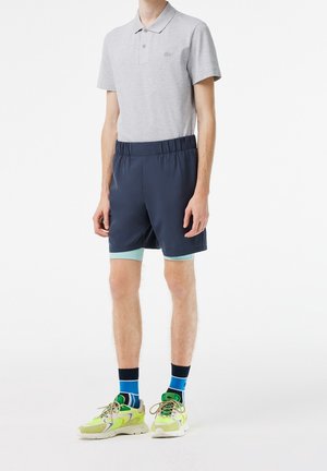 Harmaa polo paita lyhyillä hihoilla, tummansiniset shortsit joustavyöhön, ja kirkkaan vihreät lenkkarit. Siniset ja mustat kuviolliset sukat näkyvät.