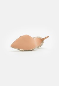 Nude high-heeled shoe with a pointed toe, adorned with glittery gold trim around the sole and a smooth, textured upper.