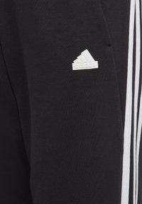 Zwarte sportbroeken van zachte stof, met witte strepen aan de zijkanten en een klein wit adidas-logo op de bovenbenen.