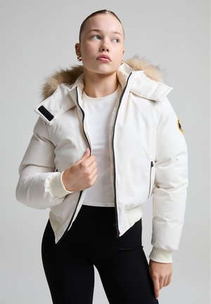 Jeune femme aux cheveux tirés en arrière porte une veste blanche à capuche en fourrure, une chemise blanche et un pantalon noir, regardant sur le côté avec une expression neutre.