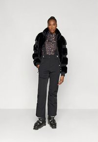 Zwarte jas van imitatiebont met gestreepte textuur, blouse met luipaardprint en zwarte broek. Gecombineerd met zwarte skistappers. Minimalistische achtergrond.