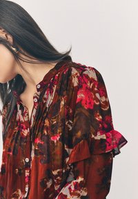 Blusa con motivo floreale rosso e marrone, con maniche arricciate e una patta con bottoni, realizzata in un tessuto leggero e strutturato.