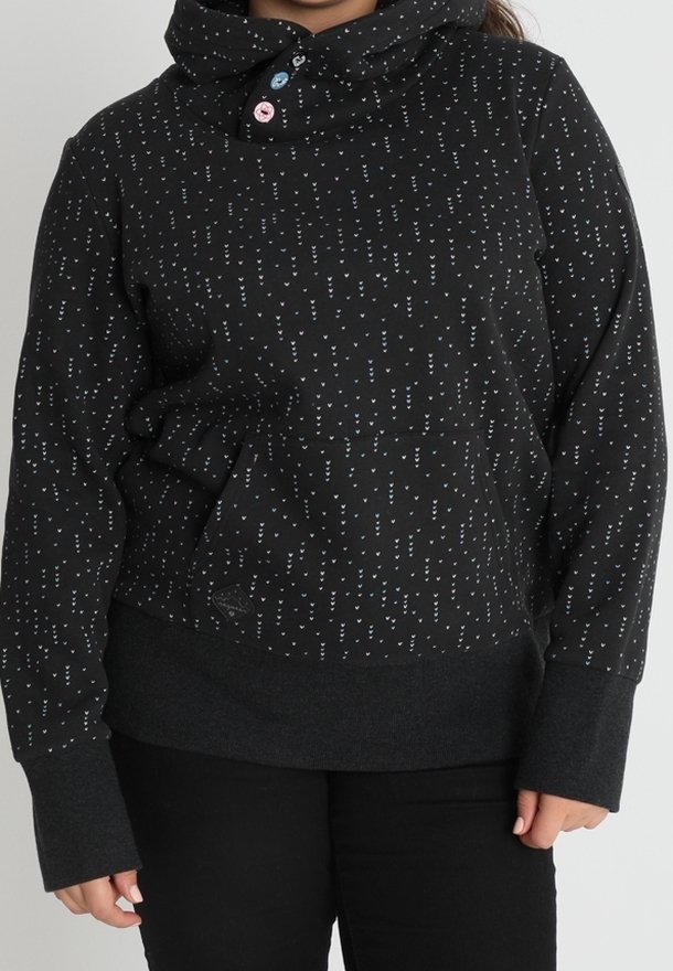 Sweat à capuche noir avec petits motifs de cœurs blancs, trois boutons colorés sur le col et larges poignets, porté avec un pantalon noir.